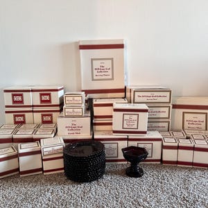May include: A collection of boxed serving dishes and plates from the 1970s Cape Cod Collection. The boxes are cream-colored with maroon accents and text. A stack of black plates and a small, dark red glass dish are also present.