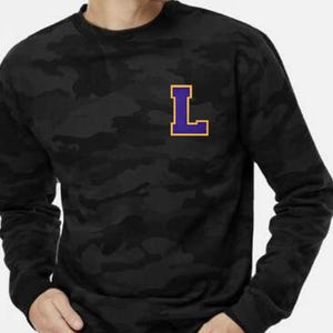 May include: Black camouflage sweatshirt with a purple and gold letter "L" patch on the chest. The long-sleeved crew neck shirt has a subtle, dark pattern throughout the fabric.