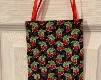 May include: A small black fabric tote bag with red handles. The bag is decorated with a repeating pattern of red cherries with green stems and leaves on a black background with white polka dots.