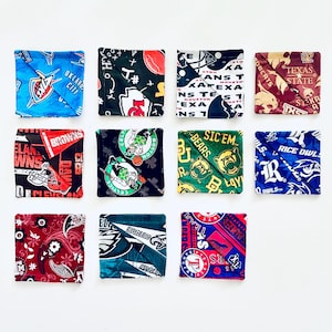 May include: Twelve square fabric coasters with various sports team logos and patterns. Designs include the Oklahoma City Thunder, Cleveland Browns, Boston Celtics, and Texas State Bobcats. Each coaster is a different color and design.