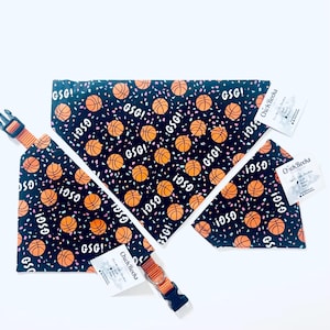 Puede incluir: Un conjunto de collar y pa&ntilde;uelo para perros con tem&aacute;tica de baloncesto. La tela negra est&aacute; estampada con balones de baloncesto naranjas, confeti y el texto "GSG!". El collar tiene una hebilla naranja y negra.
