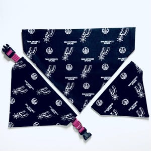 May include: Three black and white dog bandanas with the San Antonio Spurs logo and team name. The bandanas are on a white surface. One bandana is tied around a pink dog collar.