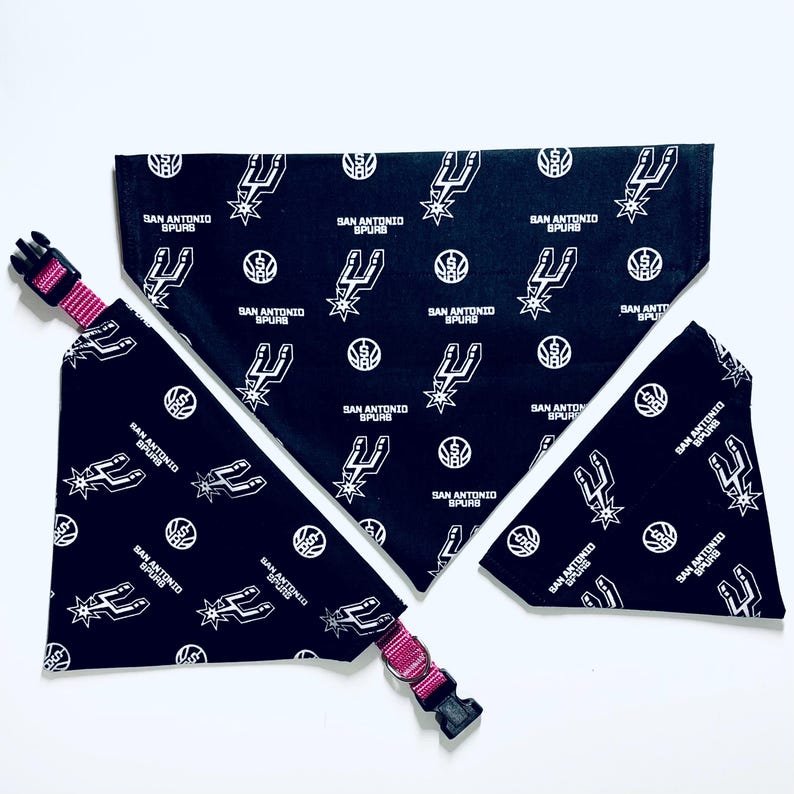 May include: Three black and white dog bandanas with the San Antonio Spurs logo and team name. The bandanas are on a white surface. One bandana is tied around a pink dog collar.