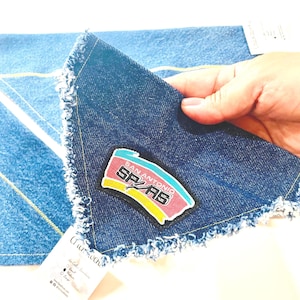 May include: A denim handkerchief with a frayed edge, featuring a San Antonio Spurs logo patch in red, black, yellow, and teal. The handkerchief is a medium wash blue denim. The corner of a second denim item is visible in the background.