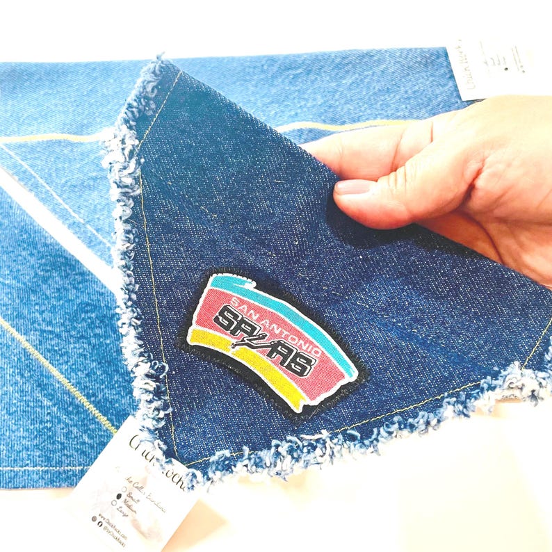 May include: A denim handkerchief with a frayed edge, featuring a San Antonio Spurs logo patch in red, black, yellow, and teal. The handkerchief is a medium wash blue denim. The corner of a second denim item is visible in the background.
