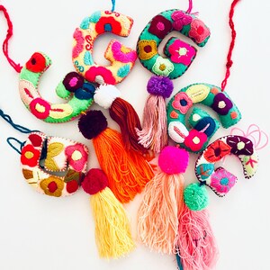 May include: Six colorful embroidered felt letter charms with pom-pom tassels. Each charm features a different letter of the alphabet and is decorated with colorful floral embroidery. The tassels are in various colors, including pink, orange, yellow, and green.