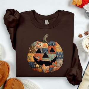 May include: Dark chocolate-colored sweatshirt featuring a patchwork pumpkin design. The pumpkin has a smiling face and is made of various floral and patterned fabrics. The sweatshirt is a crew neck style, perfect for fall and Halloween.