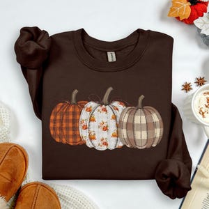 May include: Dark chocolate-colored sweatshirt featuring a trio of decorative pumpkins. The pumpkins showcase plaid, floral, and stitched patterns in orange, white, and tan. The text "Dark Chocolate" is at the bottom of the image.