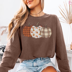 May include: A brown sweatshirt featuring three pumpkins with different patterns: plaid, floral, and solid. The word "Espresso" is printed in the upper right corner. The sweatshirt is paired with denim shorts.