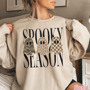 May include: A sand-colored crewneck sweatshirt featuring the black text SPOOKY SEASON in a bold, distressed font. Three ghost-shaped graphics are centered on the chest, each with a different pattern: camouflage, leopard print, and a tan checkerboard.