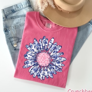 May include: A pink t-shirt with a blue and white floral design. The flower has a pink center with a leopard print pattern. The t-shirt is displayed on a white surface with a pair of blue jeans and a brown hat.