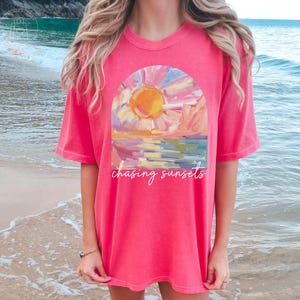 May include: A watermelon-colored t-shirt with a sunset graphic. The graphic features a colorful depiction of a sunset over water, with the words "chasing sunsets" in white script. The shirt is a relaxed fit.