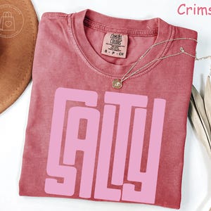 May include: Crimson-colored t-shirt with the word "SALTY" in large, pink block letters. The shirt has a crew neck and short sleeves. A silver necklace with a small pendant is draped across the shirt. The shirt is made of a soft, comfortable material.