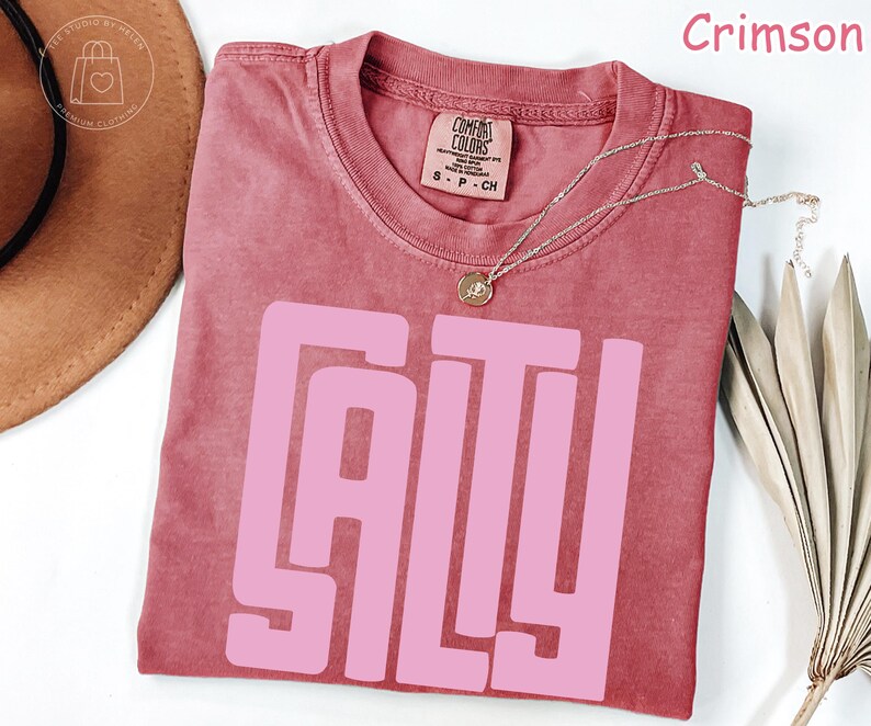May include: Crimson-colored t-shirt with the word "SALTY" in large, pink block letters. The shirt has a crew neck and short sleeves. A silver necklace with a small pendant is draped across the shirt. The shirt is made of a soft, comfortable material.