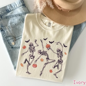 May include: Ivory-colored Comfort Colors t-shirt featuring a Halloween design. The graphic shows dancing skeletons, bats, pumpkins, and autumn leaves. The shirt is laid on top of blue jeans and a tan hat.