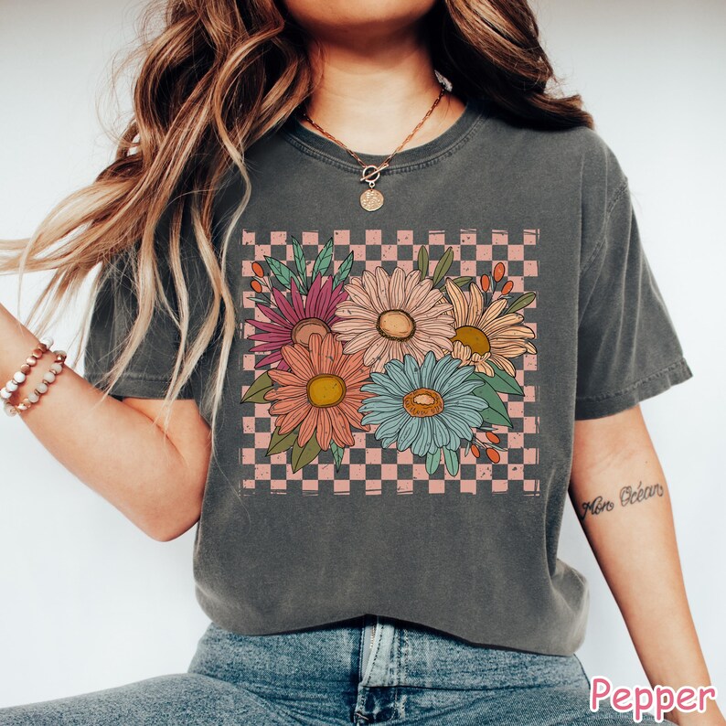 May include: Dark gray t-shirt featuring a floral design with daisies in shades of pink, orange, blue, and yellow. The flowers are set against a pink and white checkered background. The word "Pepper" is printed at the bottom.