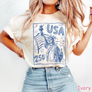Puede incluir: Una camiseta de manga corta color marfil con un estampado gr&aacute;fico azul de la Estatua de la Libertad sosteniendo una antorcha, una bandera estadounidense, estrellas y el texto USA y 250. El dise&ntilde;o est&aacute; dentro de un marco rectangular estilo boceto.