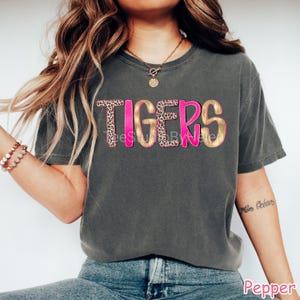 Peut inclure: Un t-shirt gris fonc&eacute; avec le mot "TIGERS" en rose et or avec un motif l&eacute;opard. Le t-shirt &agrave; manches courtes a un col rond. Le t-shirt est fait d'une mati&egrave;re douce et confortable.