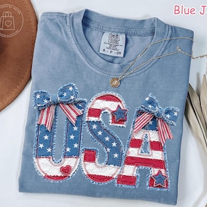 May include: A light blue t-shirt with the word "USA" appliqued in red, white, and blue denim fabric. The letters are decorated with stars, bows, and a heart. A gold necklace rests on the shirt.
