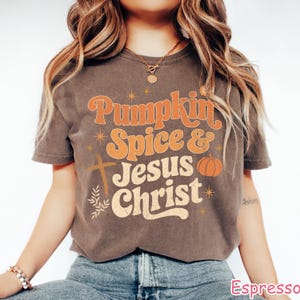Op de afbeelding: Espresso-kleurig T-shirt met de tekst "Pumpkin Spice & Jesus Christ" in oranje en cr&egrave;me letters. Het ontwerp bevat een kruis, een pompoen en steraccenten. Het shirt heeft korte mouwen en een ronde hals.