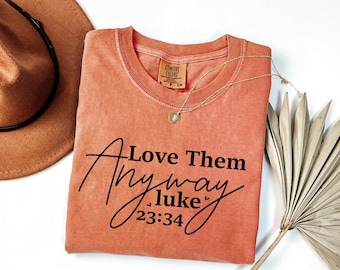 May include: A coral-colored t-shirt with the text "Love Them Anyway Luke 23:34" printed in black. The shirt is made of a soft, comfortable material.