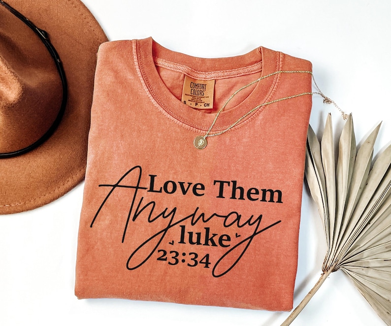 May include: A coral-colored t-shirt with the text "Love Them Anyway Luke 23:34" printed in black. The shirt is made of a soft, comfortable material.