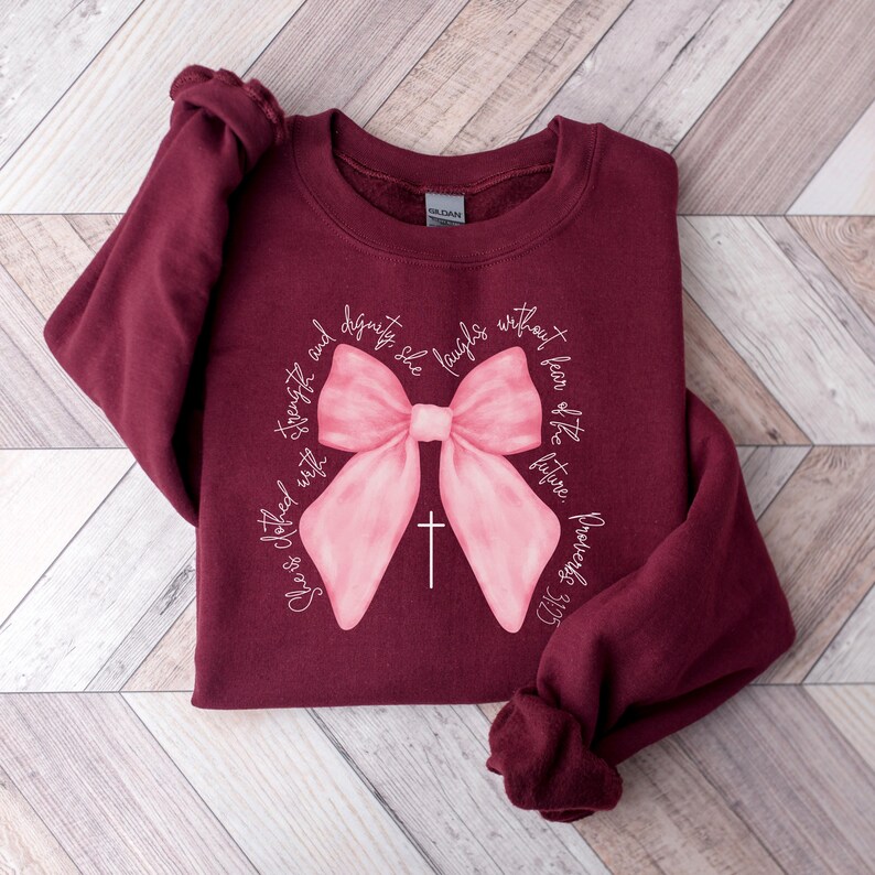 May include: Burgundy sweatshirt with a pink bow and the text "She is clothed with strength and dignity. She laughs without fear of the future. Proverbs 31:25"