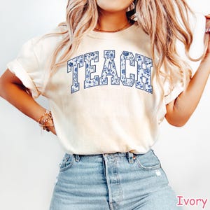 May include: A white t-shirt with a blue floral design that says "TEACH" in a varsity style font.