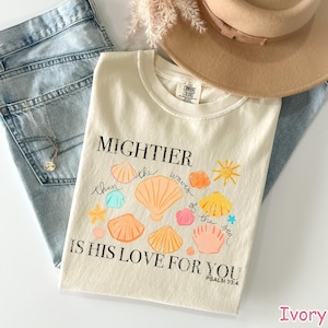 K&ouml;nnte beinhalten: Cremefarbenes T-Shirt mit dem Text "MIGHTIER than the waves of the sea IS HIS LOVE FOR YOU" und bunten Muschel-Illustrationen. Das Shirt wird mit blauen Jeans und einem beigen Hut kombiniert, f&uuml;r einen l&auml;ssigen Look.