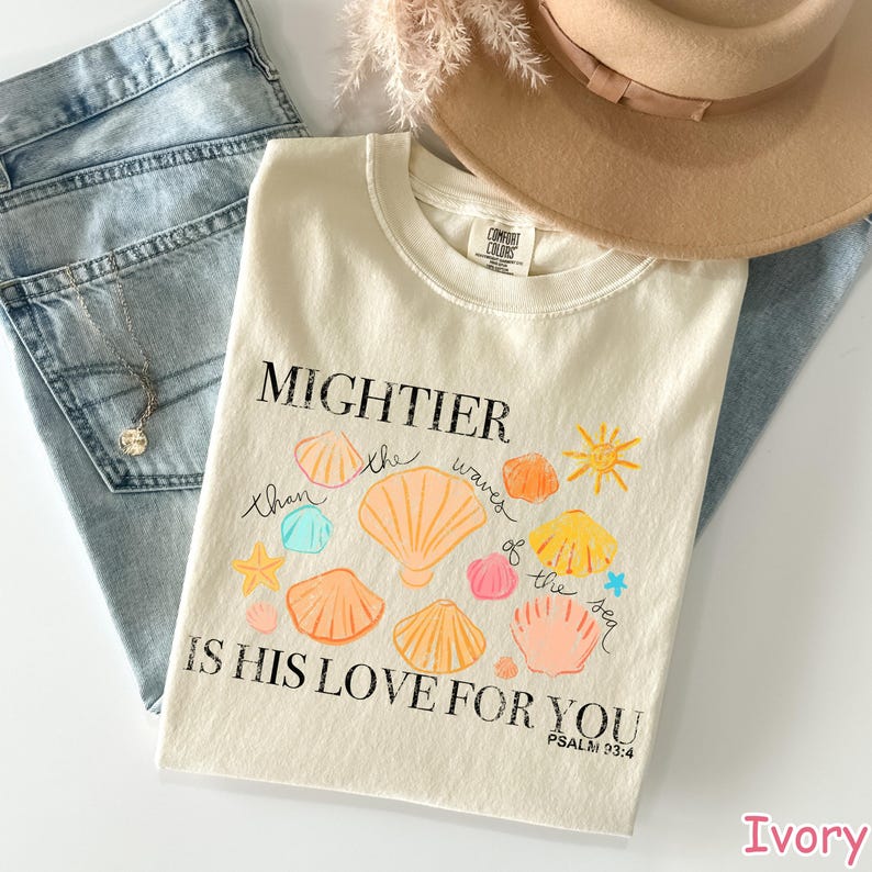 K&ouml;nnte beinhalten: Cremefarbenes T-Shirt mit dem Text "MIGHTIER than the waves of the sea IS HIS LOVE FOR YOU" und bunten Muschel-Illustrationen. Das Shirt wird mit blauen Jeans und einem beigen Hut kombiniert, f&uuml;r einen l&auml;ssigen Look.