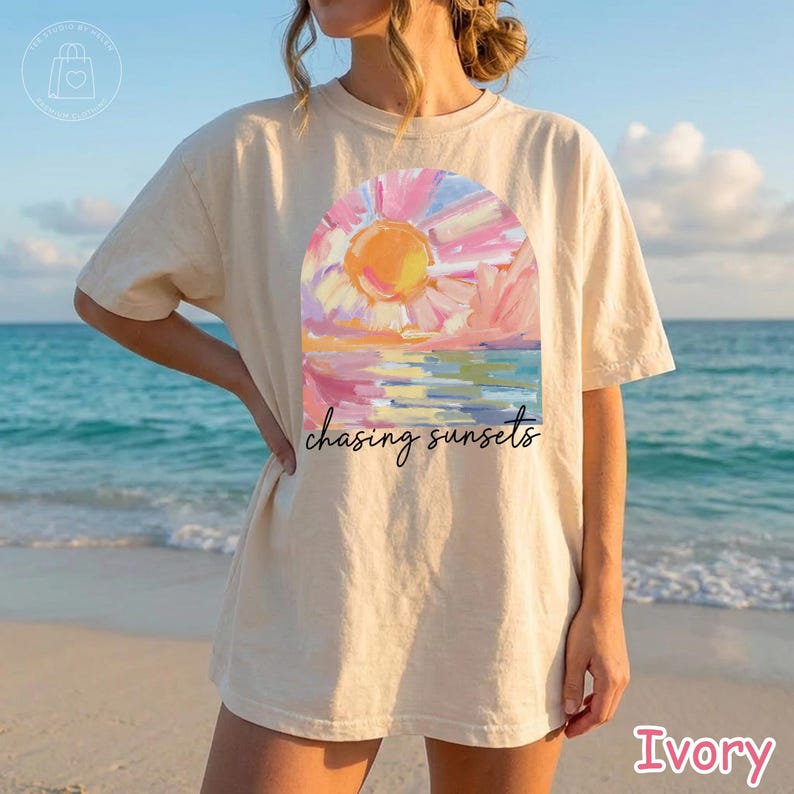 May include: Ivory-colored t-shirt featuring a colorful sunset design with the text "chasing sunsets". The artwork depicts a vibrant sun over water, with pink, orange, and blue hues. The shirt is a relaxed fit.