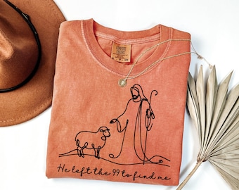 May include: A peach-colored t-shirt with a black line drawing of a shepherd and a sheep. The text "He left the 99 to find me" is printed below the drawing.