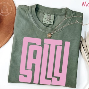 May include: A moss green t-shirt with the word "SALTY" in large pink block letters. The shirt has a crew neck and short sleeves. A gold necklace with a pendant is draped on the shirt.