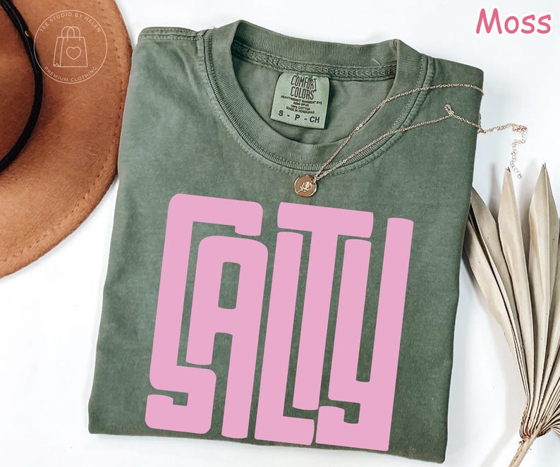 May include: A moss green t-shirt with the word "SALTY" in large pink block letters. The shirt has a crew neck and short sleeves. A gold necklace with a pendant is draped on the shirt.