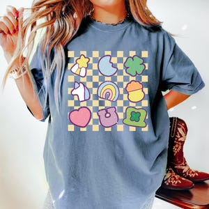 Peut inclure: Un t-shirt bleu jean &agrave; motif &agrave; carreaux avec diff&eacute;rentes images de dessins anim&eacute;s color&eacute;s, notamment un arc-en-ciel, un fer &agrave; cheval, un tr&egrave;fle &agrave; quatre feuilles, une licorne, un c&oelig;ur, une &eacute;toile, un pot d'or et un tr&egrave;fle.