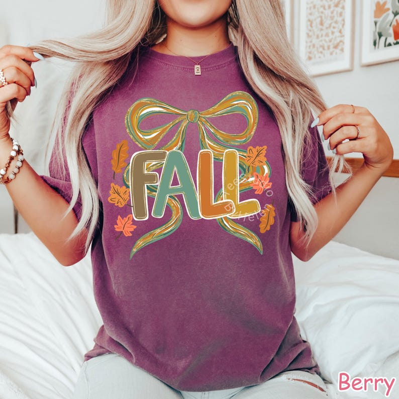 May include: Berry-colored t-shirt with a large graphic that says "FALL" in a colorful, outlined font. The design includes a bow and autumn leaves. The shirt is a short-sleeved, casual style.