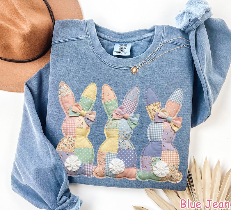May include: A blue jean sweatshirt features three appliqued patchwork bunnies with floral and gingham patterns. Each bunny has a bow and a white pom-pom tail. The sweatshirt has a crew neck and long sleeves. The text "Blue Jean" is at the bottom right.
