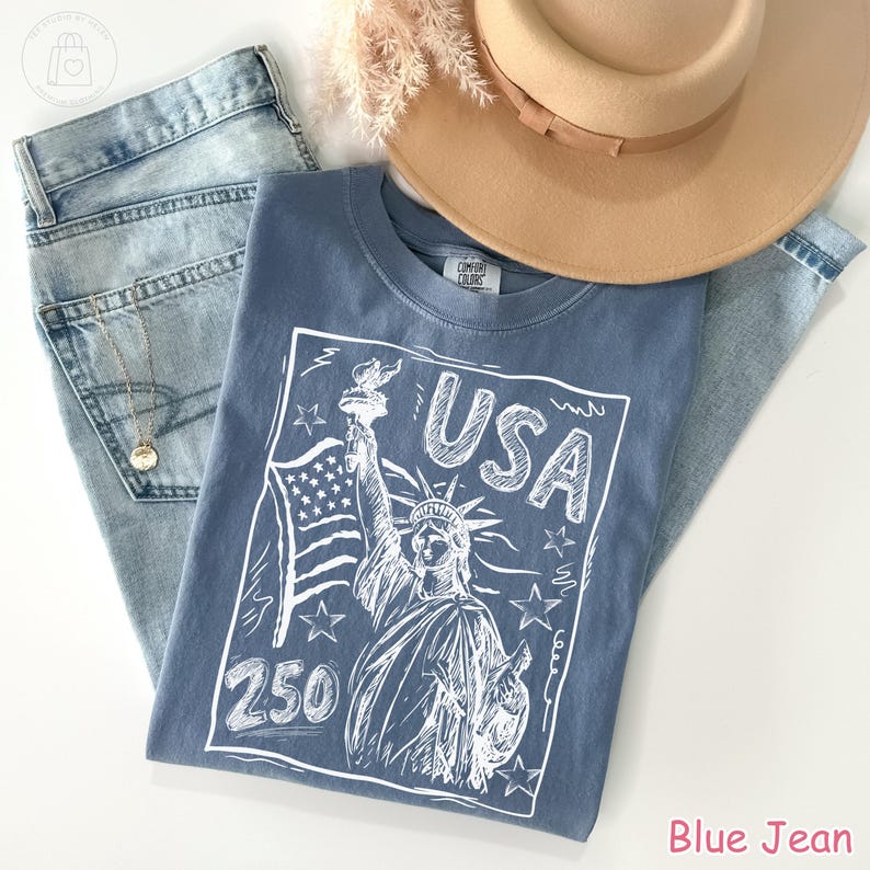 Puede incluir: Una camiseta azul con un gr&aacute;fico blanco de la Estatua de la Libertad, la bandera estadounidense, el texto USA y 250, junto a unos vaqueros azul claro, un sombrero beige de ala ancha y una fina cadena dorada con un peque&ntilde;o colgante redondo.