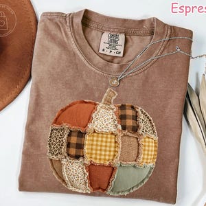 May include: An espresso-colored short-sleeve t-shirt featuring a large, central pumpkin graphic made of various fabric patches. The patches include brown and yellow gingham, leopard print, floral patterns, and solid earth tones with frayed edges.