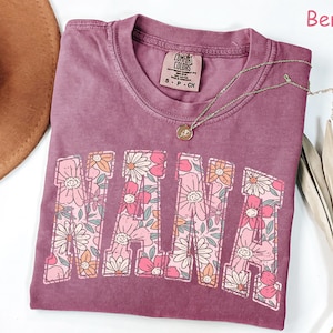 May include: A mauve colored t-shirt with the word "MAMA" printed in large letters. The letters are filled with a floral pattern in pink, orange, and green. The shirt has a distressed look and is made of a soft, comfortable fabric. The text "COMFORT COLORS" is printed on a small tag attached to the shirt.