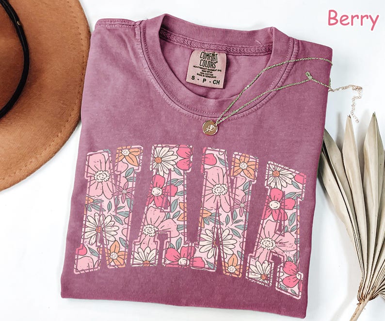 May include: A mauve colored t-shirt with the word "MAMA" printed in large letters. The letters are filled with a floral pattern in pink, orange, and green. The shirt has a distressed look and is made of a soft, comfortable fabric. The text "COMFORT COLORS" is printed on a small tag attached to the shirt.