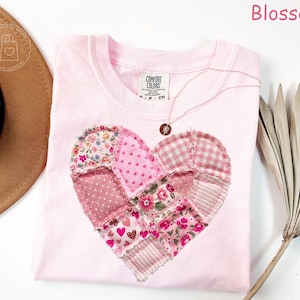 May include: A light pink t-shirt with a patchwork heart design. The heart is made of various pink fabrics with floral and polka dot patterns. A delicate necklace with a pendant rests on the shirt. The word "Blossom" is in pink text.