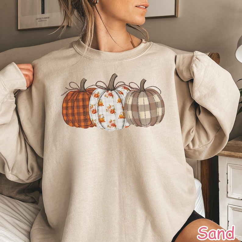 Puede incluir: Sudadera beige con tres calabazas decorativas. Las calabazas tienen diferentes patrones: cuadros naranjas, blanco con detalles florales y cuadros marrones. Cada calabaza tiene un tallo marr&oacute;n y un lazo decorativo.