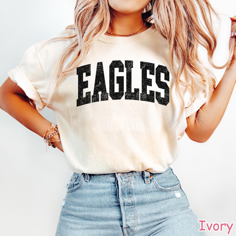 May include: Ivory-coloured t-shirt featuring the word "EAGLES" in bold, black, distressed lettering. The shirt is a short-sleeved crew neck style. The word "Ivory" is printed in pink at the bottom right corner.
