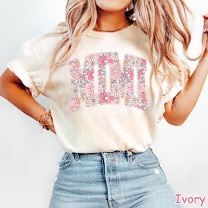 K&ouml;nnte beinhalten: Elfenbeinfarbenes T-Shirt mit dem Wort "MOM" in einem floralen Design. Die Buchstaben sind mit rosa, wei&szlig;en und pfirsichfarbenen Blumen gef&uuml;llt. Das Shirt hat eine lockere Passform und kurze &Auml;rmel.