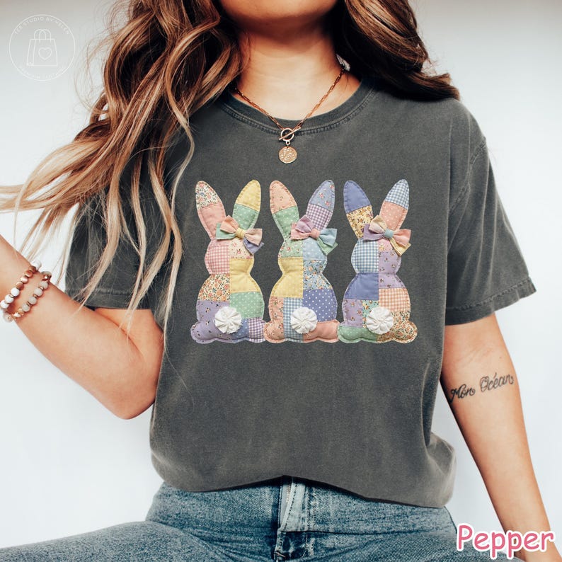 K&ouml;nnte beinhalten: Dunkelgraues T-Shirt mit drei Patchwork-Hasen-Designs. Die Hasen haben bunte Muster und Schleifen. Die Muster umfassen Blumen-, Tupfen- und Karomuster. Das Wort "Pepper" steht unten rechts.