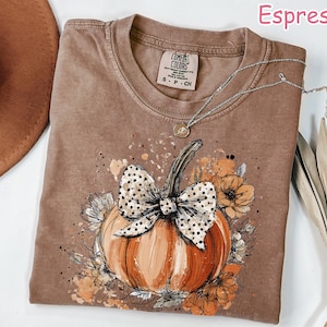 May include: Espresso-colored t-shirt featuring a pumpkin design with a polka-dot bow, surrounded by flowers. The shirt has a crew neck and short sleeves. A silver necklace with a pendant is draped on the shirt. The shirt is labeled "Comfort Colors".