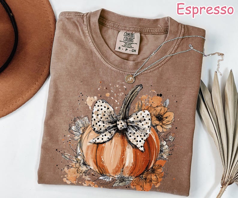 May include: Espresso-colored t-shirt featuring a pumpkin design with a polka-dot bow, surrounded by flowers. The shirt has a crew neck and short sleeves. A silver necklace with a pendant is draped on the shirt. The shirt is labeled "Comfort Colors".