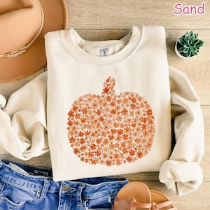 May include: A sand-colored sweatshirt featuring a pumpkin design made of orange and peach-colored flowers. The long-sleeved top is made of a soft material and is perfect for fall. The word "Sand" is printed in the upper right corner.