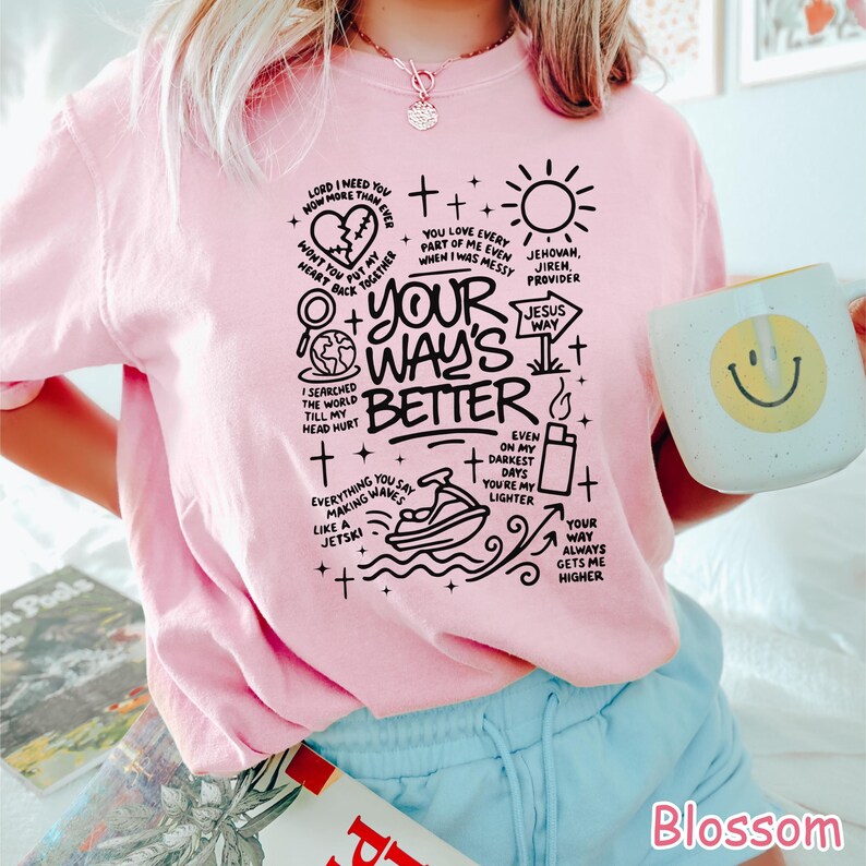 Op de afbeelding: Een lichtroze sweatshirt met een zwart grafisch ontwerp met de tekst "Your Way's Better" en andere zinnen en illustraties. Een witte mok met een gele smiley wordt in de hand gehouden.